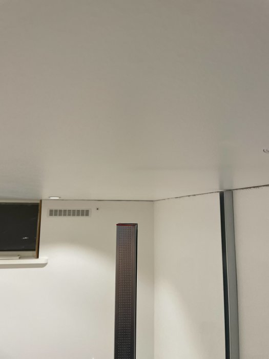 Ett hörn av ett rum med en golvstående högtalare, väggventilation och en del av en tavla.