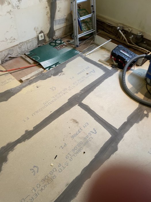 Renoveringsarbete på golv, isoleringsmaterial synligt, verktyg och ledningar, inomhusbyggnadsscenario, delvis oavslutat projekt.