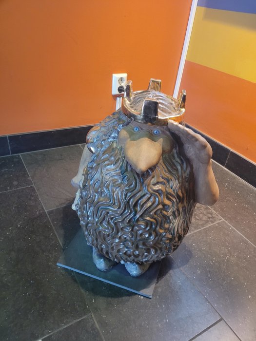 En skulptur föreställande en djurlignande figur med krona, mot en orange-gul vägg och mörkt golv.