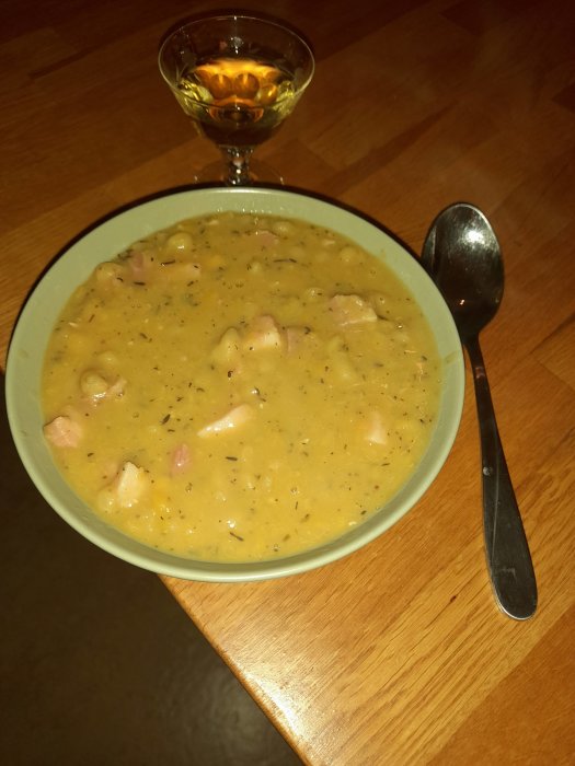En skål med grönaktig soppa, möjligtvis ärtsoppa, sked bredvid, ett glas dryck ovanför på ett träbord.