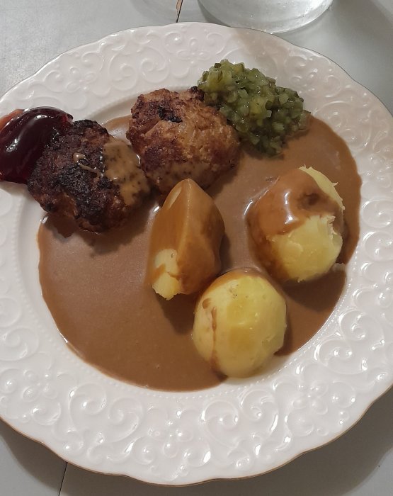 Maträtt med köttbullar, potatis, brunsås, lingonsylt och pressgurka på dekorerad tallrik.
