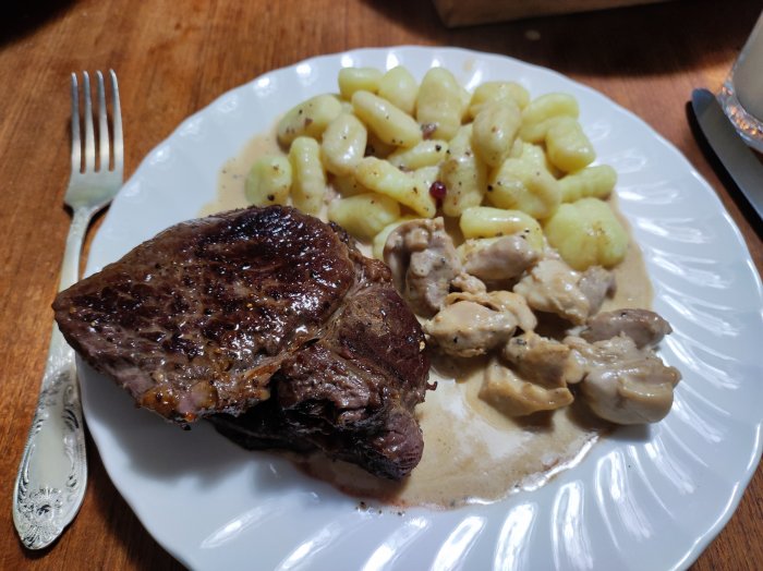 Grillad biff, svampsås, gnocchi på tallrik med gaffel, träbord. Måltid serverad, tilltalande matpresentation.