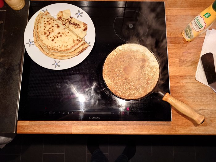 Ett kök med spis, stekpanna och pannkakor på en tallrik. Matlagningsolja och hushållspapper synliga.