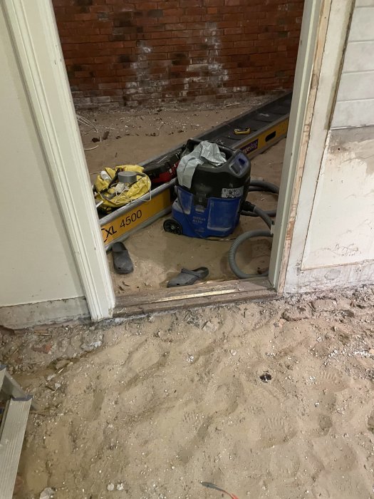 Renoveringsplats med röd tegelvägg, konstruktionsdammsugare, kratta, nivelleringsverktyg och spridd byggavfall.