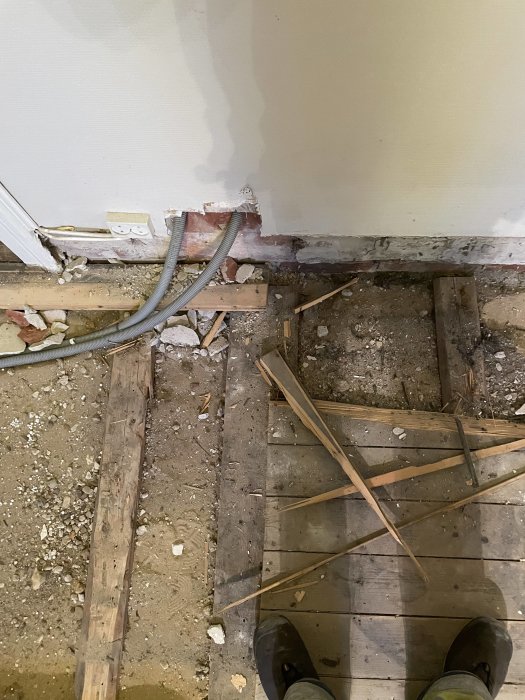Renoveringsarbete, avrivet golv, träbjälkar, kabel, spillror, öppen vägg, byggarbetsplats, stående persons skodon synliga.