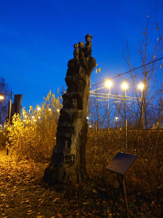 Träskulptur med figurer, kvällshimmel, gatljus, skylt, utanför i skymning.