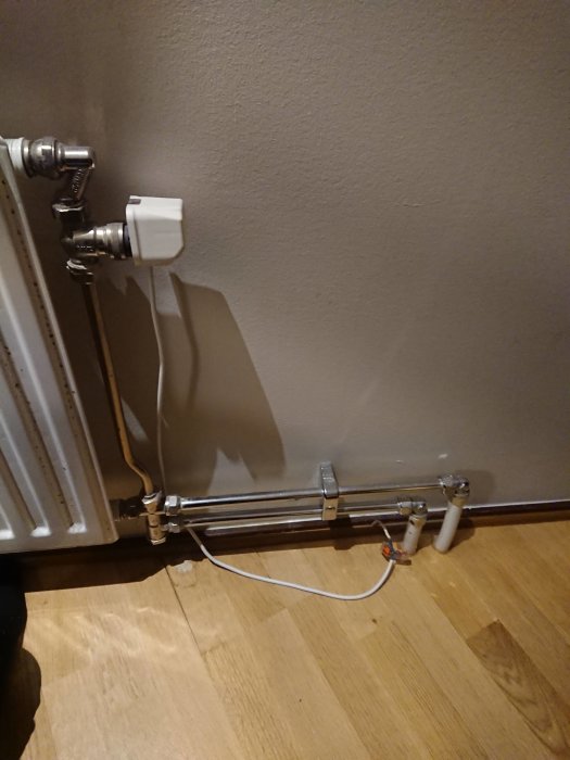 Radiator med termostat, rör på vägg, eluttag, trägolv, skuggor, vit sladd, inomhus, ingen person.