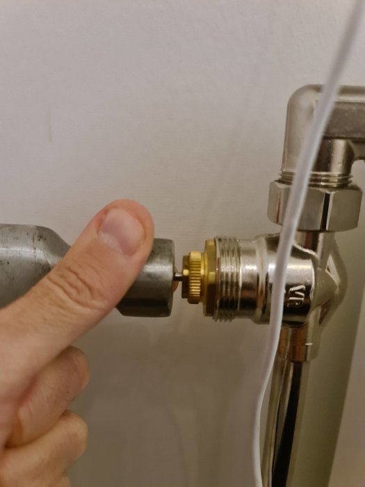 Finger pekar på termostatventil på radiator. Anslutning, rör, vägg i bakgrunden.