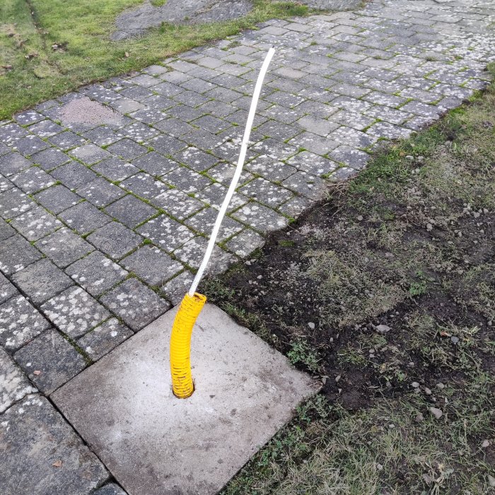 En gul snöskyffel lutar mot en trottoar av betongplattor och gatsten, med gräs och jord intill.