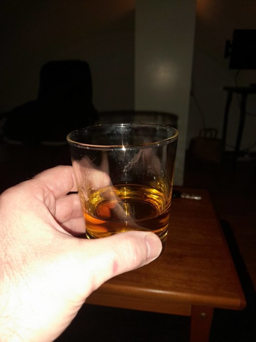 Hand håller whiskyglas, mörkt rum, trämöbel i bakgrunden.