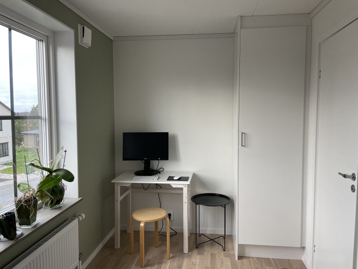 Minimalistisk arbetsrum med skrivbord, skärm, stol, pall och växter vid fönster, enkel och funktionell design.