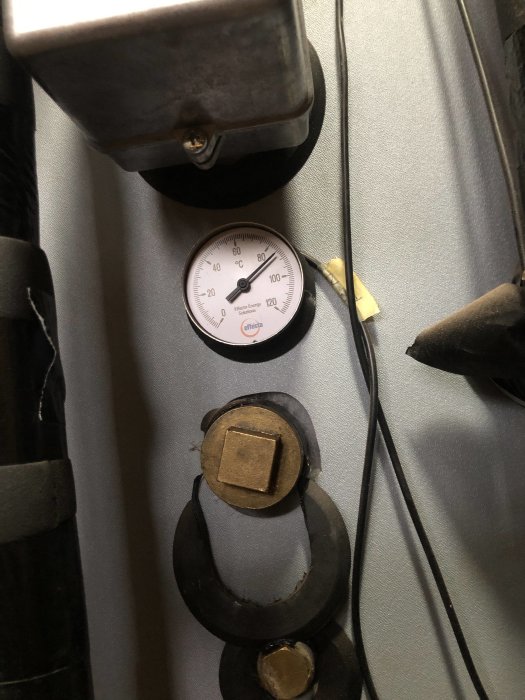 Termometer visar cirka 60 grader Celsius, industriella rör och komponenter, metallstrukturer, kablar, skuggat område.