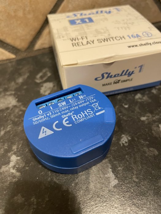 Blå Wi-Fi-reläbrytare, Shelly 1, CE-märkt, elektronikkomponent, relaterad låda i bakgrunden.