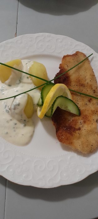 Panerad fiskfilé, kokt potatis, dillsås, citron, gurka på vitt dekorerat fat.