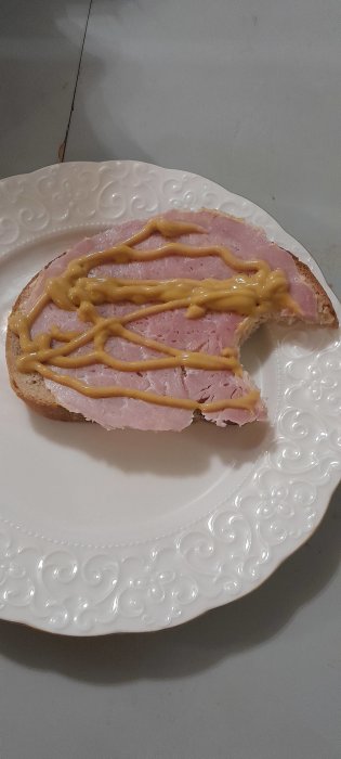 En skiva bröd med skinka och senap på ett vitt dekorerat fat.