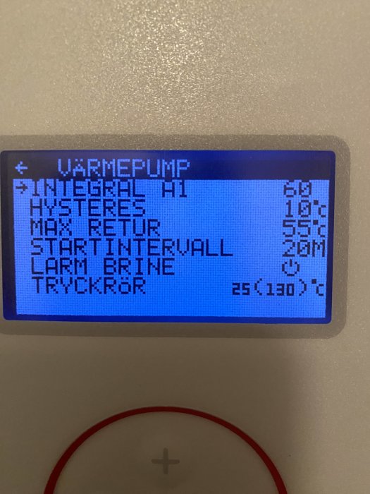 Digital display för värmepump med text och siffror som visar temperatur och inställningar, omgivet av vit vägg.