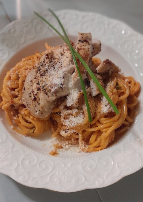 Pasta med tomatsås, strimlad kyckling, parmesan, gräslök på dekorerat vitt fat.