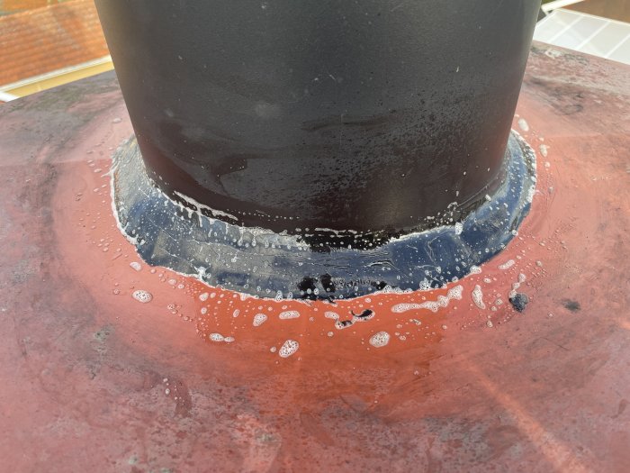 Svart rör på röd yta, vatten och bubblor runt tätning, våt yta, takgenomföring med tätningsmassa.