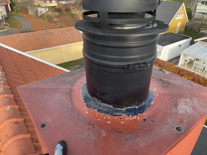 Skorsten på tak med roterande ventil, tegelpannor, bostadshus i bakgrunden, soligt väder, del av sko synlig.