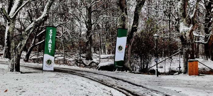 Vinterscen med "START"-banderoll, snötäckt väg och träd, skyltar för orienteringsklubb.