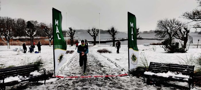 Park i vinterskrud, person löper målområde, snöig dag, folksamling i bakgrunden, kal träd, grå himmel.