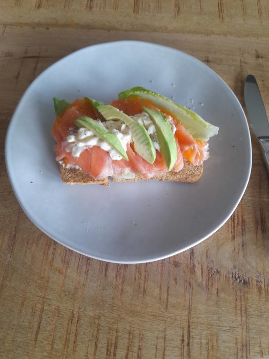 Ett stycke rostat bröd med lax, avokado, gräddfil och sallad på vit tallrik.