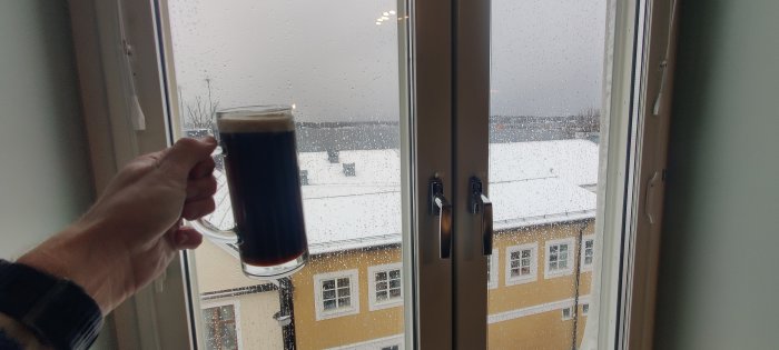 Hand håller mörkt ölglas framför fönster, regndroppar, snötäckt gatuvy, gul byggnad, disigt väder.