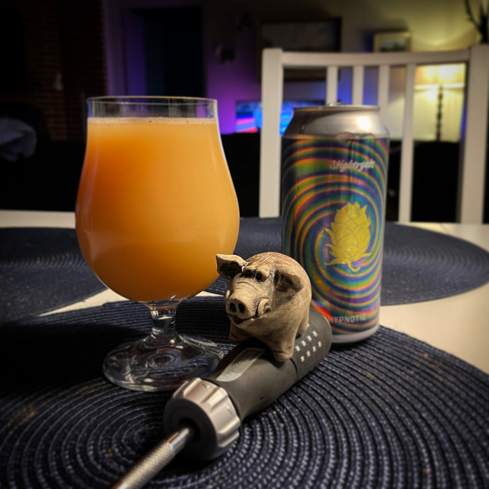 Ett glas med orange dryck, en färgglad ölburk, och en liten grisfigur på en blå duk.