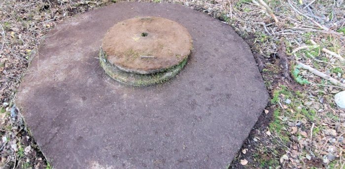 Rund betongkonstruktion med mossa, troligtvis en avlopps- eller inspektionsbrunn, inbäddad i mark med löv och kvistar.