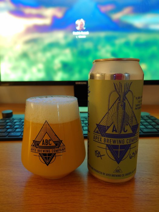 Ölglas med skum vid dator, ölburk med etikett, Apex Brewing Company, inomhus.