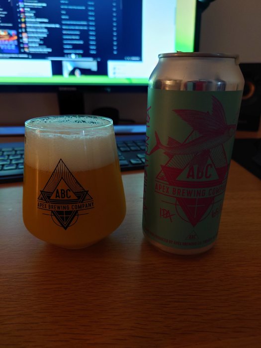 Ölglas med skum intill burk, Apex Brewing logotyp, datorskärm med kod i bakgrunden.