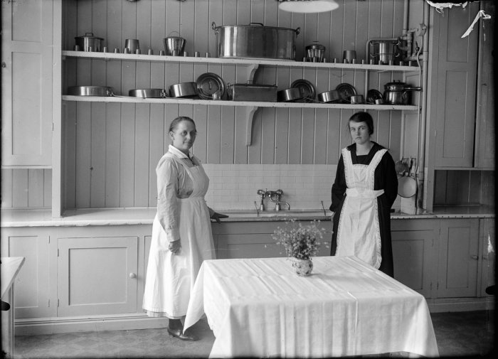 Två kvinnor i förkläden står i ett gammalt kök med köksredskap och blomvas på bordet. Svartvit bild.