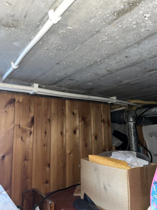 Cluttered attic or crawlspace with wooden walls, exposed pipes, storage boxes, and low lighting.