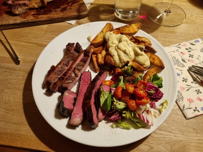 Grillad biff, kryddiga potatisklyftor, bearnaisesås, blandad grönsallad, cocktailtomater, vattenglas, vin, träskärbräda.