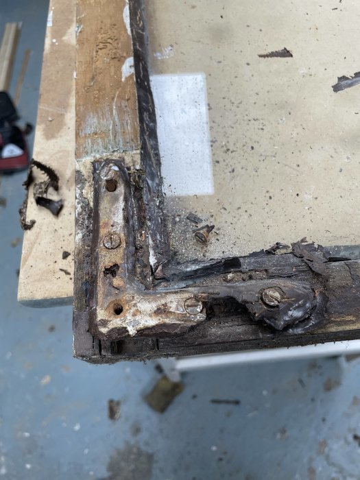 Närbild på rostig metallscharnier och delar på träyta med spån och skruvar. Slitage och korrosion syns tydligt.