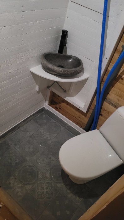 Ett ofullständigt badrum med toalett, handfat, blåa rör och trägolv. Under konstruktion eller renovering.