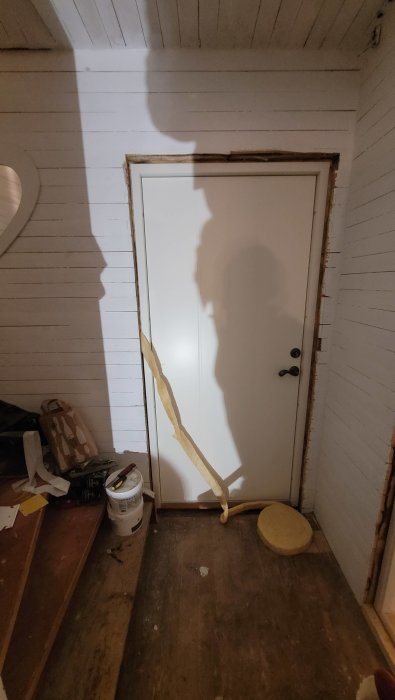 En vit dörr med skugga av människa, byggmaterial och verktyg på ett byggarbetsområde inomhus.