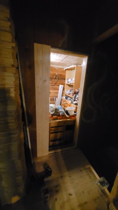 Utsikt mot en rörig verkstad genom en öppen dörr, träväggar, skymd belysning, byggmaterial och verktyg syns.