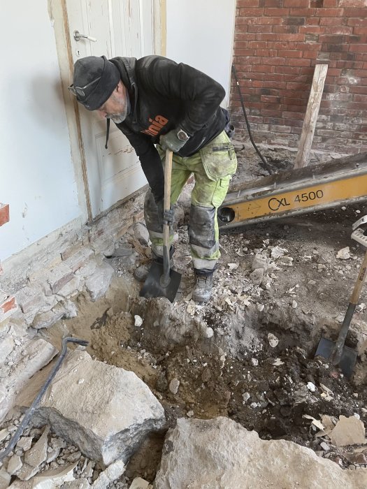 Arbetare gräver i rasmassor inne i en byggnad, nära en tegelvägg och träbjälkar.