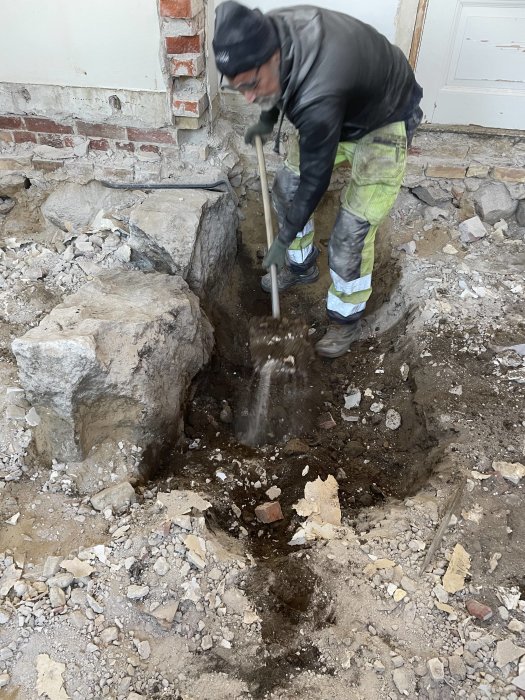 Arbetare gräver i mark, sten, grus och jord nära byggnadsvägg, arbetskläder, verktyg, rör i syn.