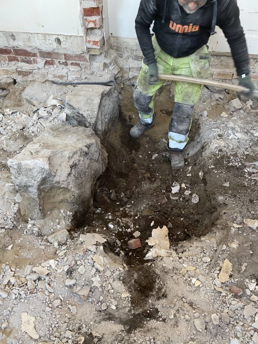 Person gräver i sönderbruten betonggolv inne i byggnad, arbetskläder och verktyg synliga.