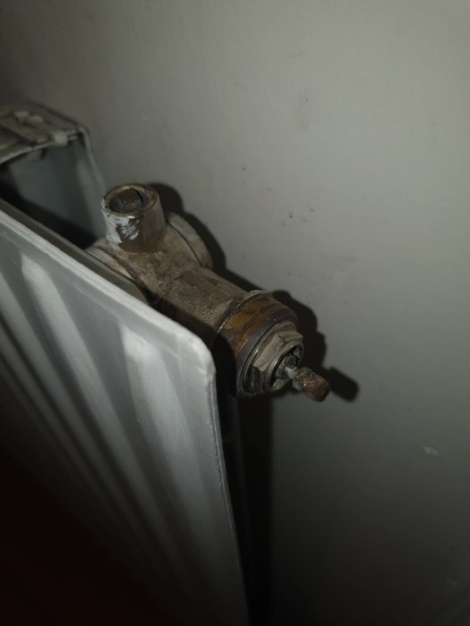 Närbild på rostig termostatventil på vit radiator, mot grå vägg.