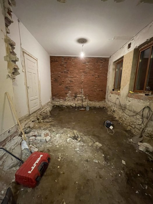 Ett rum under renovering med utsatt tegelvägg, grusig golv, och byggverktyg.