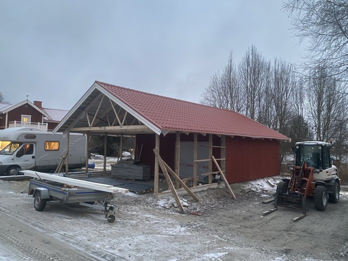 Byggarbetsplats med rött hustak under konstruktion, traktor, släpvagn, husvagn och snöfläckar.