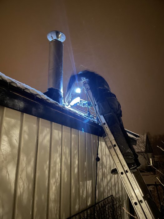 Person arbetar på tak vid skorsten på kvällen under snöfall med en stege.