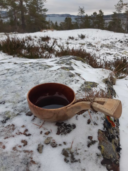 En träkåsa fylld med dryck, snöfläckad mark, skog och sjö i bakgrunden, halvmulet väder.