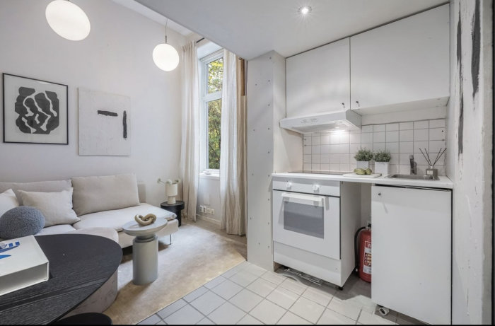 Liten, modern lägenhet med kombinerat vardagsrum och kök, vita väggar, minimalistisk inredning, konst, fönster.
