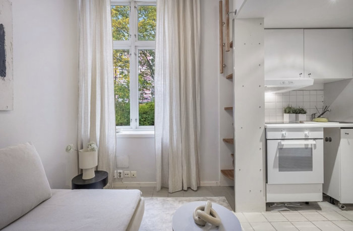 Ljus vardagsrum med kökshörn, stora fönster, grönska utsikt, vit minimalistisk inredning, trädetaljer, konstväxt.