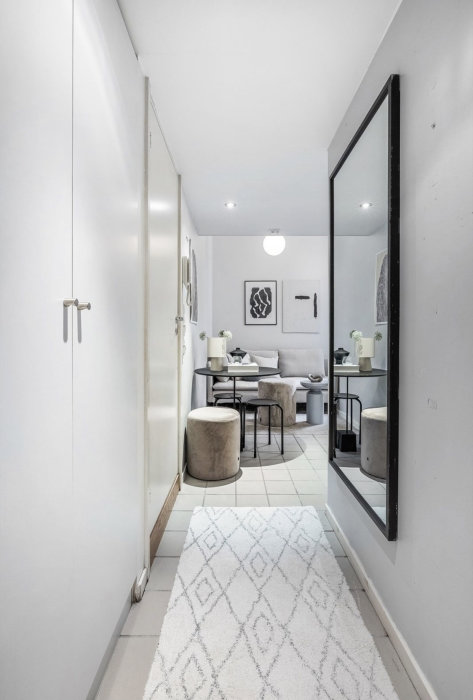 Smal hall med vit matta, spegelvägg, leder till ljust vardagsrumsmöblerat område. Modern och minimalistisk stil.