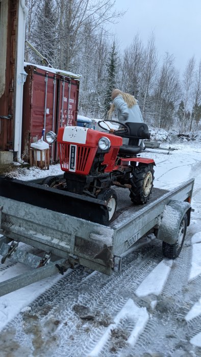 Person lastar röd trädgårdstraktor på släpkärra i snöigt landskap.